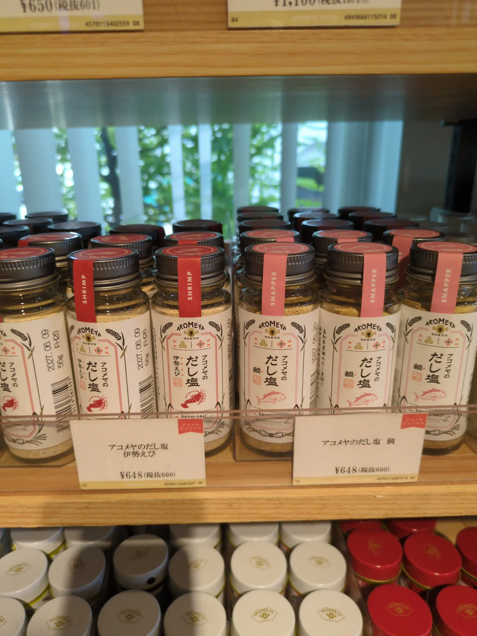 木の棚の一段に、丸い透明ガラスの瓶が左右二群に分けて、それぞれ何列も奥へ並んでいる。ラベルはどちらも同じ書式で、上にAKOMEYA TOKYOのロゴと稲穂、中央に縦書きの「アコメヤのだし塩」、その脇に赤い角印、下に赤い線画、最下部に欧文のDASHI SALT。左の群はキャップに濃い赤の帯で「SHRIMP」、線画はえび。右の群はサーモンピンクの帯で「SNAPPER」、線画は鯛。手前の値札はどちらも648円(税抜600)