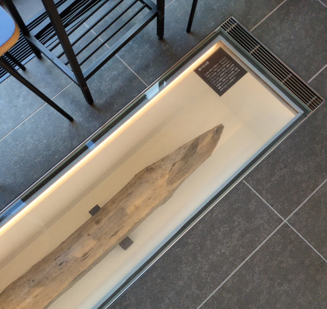 同じ床の展示ケースを別角度から見たところ。松の丸太杭の、削って尖らせた先端が画面の奥に向いている。杭の表面は淡い茶色で、木目に沿って割れが走り、黒い染みが点々と乗っている。ケースの右奥に小さな茶色の説明板が上を向けて置かれている