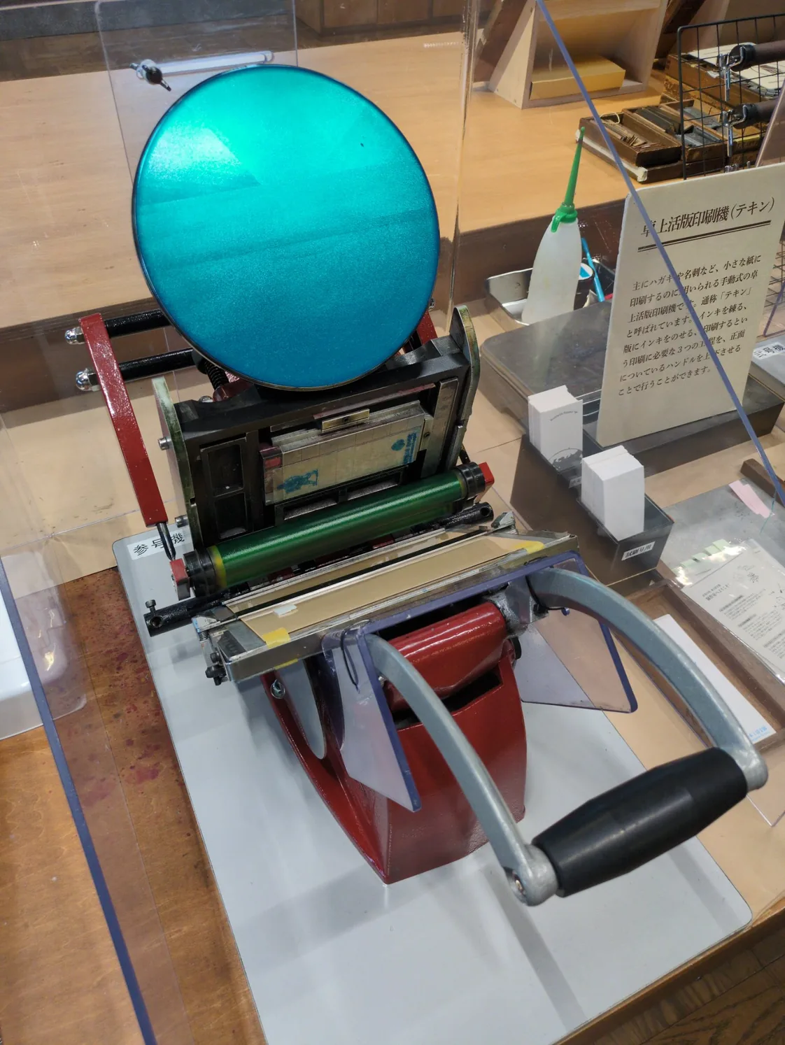 アクリルケース越しに見た卓上活版印刷機(テキン)。機体の左側に白い縦書きの札で「参号機」。いちばん上に円いインキ練り盤があり、練られているのはターコイズのインキ。その下に版を挟んだ黒い枠、中央に緑のローラー、下に赤い胴、手前に黒いグリップの大きな操作ハンドルが一本だけ突き出している。右にはこの機械の解説板と、試し刷り用の白と桃色の紙が置いてある